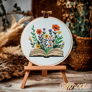 May include: A cross-stitch embroidery hoop featuring a colorful floral design emerging from an open book. The flowers include orange poppies, yellow daisies, and purple blooms. The embroidery is displayed on a small wooden easel.