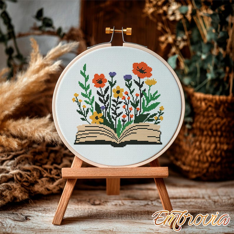 May include: A cross-stitch embroidery hoop featuring a colorful floral design emerging from an open book. The flowers include orange poppies, yellow daisies, and purple blooms. The embroidery is displayed on a small wooden easel.