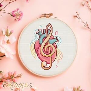 Könnte beinhalten: Ein Kreuzstich-Stickrahmen mit einem rosa Herzen mit türkisfarbenen anatomischen Details und einem goldenen Violinschlüssel. Das Design umfasst Noten und wellenförmige Linien in Gelb und Türkis. Der Rahmen befindet sich vor einem rosa Hintergrund mit floralen Akzenten.
