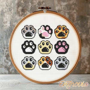 May include: A cross-stitch embroidery hoop with nine colourful paw prints. The paw prints are in various colours, including black, grey, yellow, and pink, arranged in a grid. The wooden embroidery hoop has the word "Embrovia" in the bottom right corner.