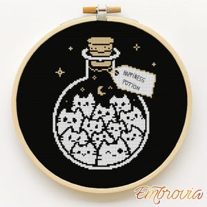 Puede incluir: Arte de punto de cruz en un aro de madera. El diseño presenta un fondo negro con una botella de poción blanca llena de caras de gato. La botella tiene un tapón de corcho y una etiqueta que dice "Happiness Potion."