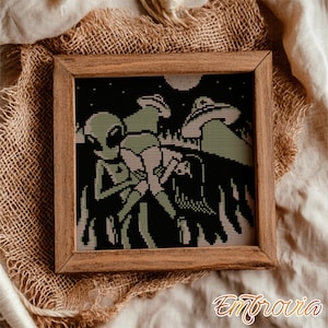 May include: A framed cross-stitch artwork featuring a retro-style alien abduction scene. The design uses black, green, and tan colors. The artwork is set against a dark night sky with a full moon and stars. The frame is made of wood.
