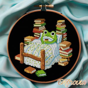 May include: Cross-stitch embroidery in a wooden hoop features a frog in bed surrounded by stacks of books. The frog is green with a white and blue patterned blanket. The background is black.