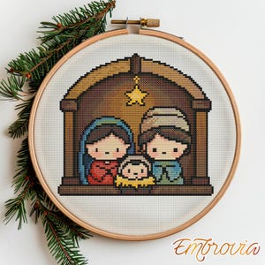 May include: A cross-stitch embroidery of a Nativity scene, featuring Mary, Joseph, and baby Jesus in a stable. The design is framed in a wooden hoop, with a star above the stable. Green pine branches are in the background.