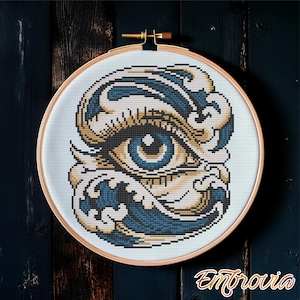 May include: A cross-stitch embroidery in a wooden hoop features a detailed eye design with blue, tan, and white colors. The eye is surrounded by stylized waves. The embroidery is set against a dark wood background. The word "Embrovia" is in the bottom right corner.