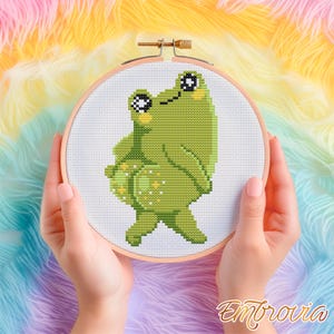 Può includere: Un telaio da ricamo a punto croce con un disegno di rana verde. La rana ha grandi occhi ed è ambientata su uno sfondo bianco. Il telaio da ricamo è tenuto in mano, con uno sfondo di filato colorato. Il testo "Embrovia" si trova nell'angolo in basso a destra.