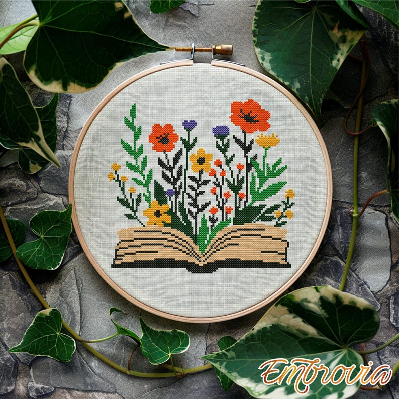 May include: A cross-stitch embroidery hoop featuring an open book with colorful flowers. The flowers include orange poppies, yellow and purple blooms, and green foliage. The embroidery is set against a light fabric, framed by a wooden hoop.