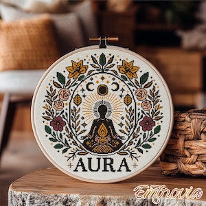 May include: A round cross-stitch embroidery hoop featuring a detailed design with the word "AURA". The design includes a central figure in a meditative pose, surrounded by floral and celestial elements in shades of gold, green, pink, and black.