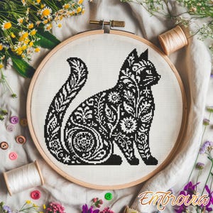 May include: A black and white cross-stitch cat design in a wooden embroidery hoop. The cat is filled with floral and leaf patterns. The word "Embrovia" is in the bottom right corner. The background includes flowers, thread spools, and buttons.