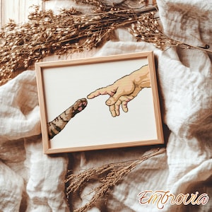 Könnte beinhalten: Ein gerahmtes Kreuzstichbild, das eine menschliche Hand zeigt, die sich einer Katzenpfote nähert, in Anlehnung an Michelangelos 'Erschaffung Adams'. Das Kunstwerk verwendet Beige-, Braun- und Weißtöne in einem hellbraunen Holzrahmen. Der Text "Embrovia" ist enthalten.