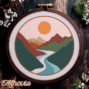 Puede incluir: Un patrón de bordado de punto de cruz de un paisaje con un río serpenteando a través de montañas y un gran sol naranja en el cielo. Los colores son rosa, naranja, amarillo, marrón, verde y azul.
