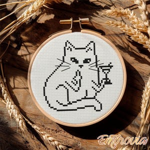 May include: A cross-stitch embroidery hoop featuring a black and white cat holding a martini glass. The cat is sitting and has a sassy expression. The embroidery is set in a wooden hoop, with wheat stalks in the background.