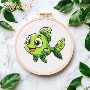 May include: A green cartoon fish with big eyes and a smiling mouth is embroidered on white fabric in a wooden embroidery hoop.