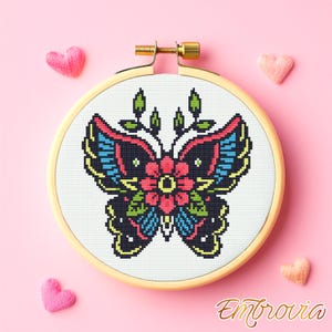 Puede incluir: Un diseño de mariposa bordada en punto de cruz en un bastidor de madera. La mariposa tiene un cuerpo negro, alas rojas, azules y amarillas, y una flor central. El fondo es blanco, y el bastidor está sobre un fondo rosa con decoraciones en forma de corazón.