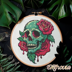 May include: Cross-stitch embroidery of a teal skull with red roses and green leaves, set in a wooden hoop. The design features a detailed pixelated style, with the word "Embrovia" in a cursive font at the bottom.