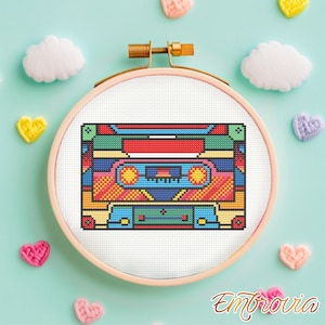 May include: A colorful cross-stitch design of a retro cassette tape, framed in a wooden embroidery hoop. The cassette tape features a vibrant mix of red, blue, green, and yellow. The background includes decorative hearts and clouds.