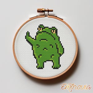 May include: A cross-stitch embroidery hoop featuring a green frog with a raised arm. The frog is made of small, square stitches. The embroidery hoop is round, with a wooden frame and a brass screw. The text "Embrovia" is in the bottom right corner.