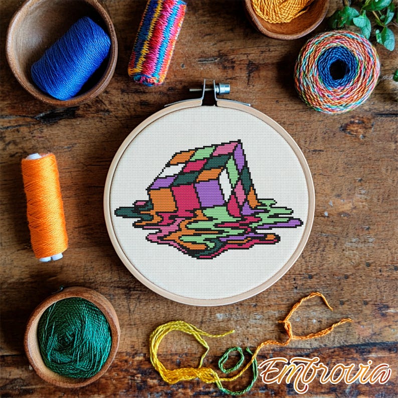 May include: A cross-stitch embroidery hoop featuring a colorful, melting Rubik's Cube design. The artwork includes shades of pink, purple, green, and orange. Various spools of thread and yarn in complementary colors surround the hoop on a wooden surface.