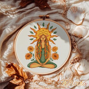 Peut inclure: Broderie au point de croix dans un cercle en bois. Elle représente une femme méditant sous un soleil, entourée de tournesols. La femme a de longs cheveux roux et porte une robe verte. Idéal pour la décoration intérieure.
