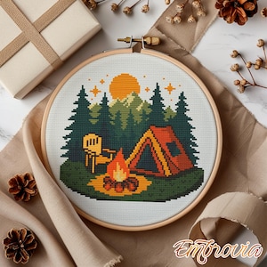 May include: A cross-stitch embroidery hoop featuring a camping scene. The design includes a tent, campfire, trees, and a setting sun. The color palette consists of orange, green, yellow, and brown. The embroidery is set in a wooden hoop.