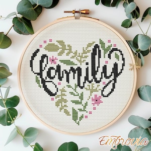 Op de afbeelding: Een borduurring met kruissteek met het woord "Family" in zwart schrift, omringd door een hartvormig ontwerp van groene bladeren en roze bloemen. De borduursel staat op een witte stof, omlijst door een houten ring.