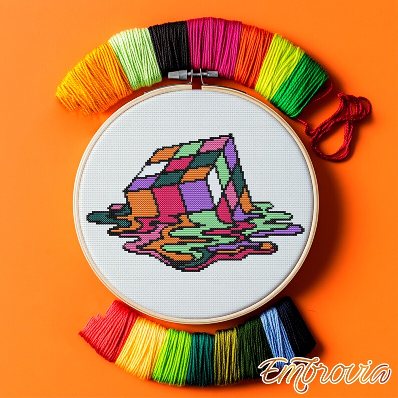 May include: A cross-stitch embroidery hoop featuring a colorful, melting cube design. The artwork uses various colors, including pink, purple, green, and orange. Bundles of colorful embroidery floss surround the hoop, with the word "Embrovia" in the bottom right corner.