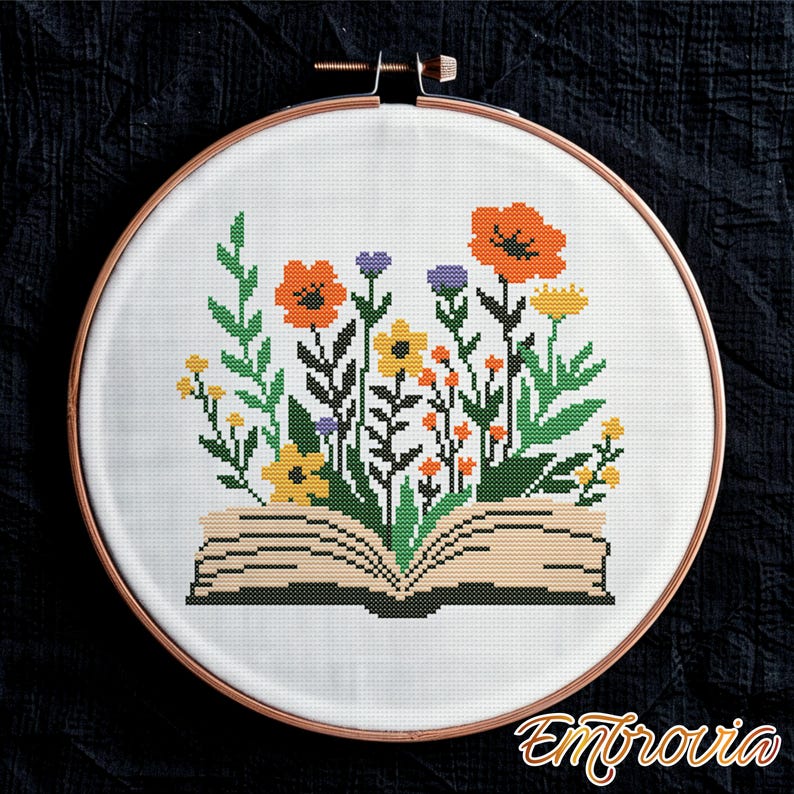 May include: Cross-stitch embroidery of a book with colorful flowers. The open book features orange poppies, yellow and purple flowers, and green leaves. The embroidery is set in a wooden hoop against a black background. The word "Embrovia" is in the bottom right corner.