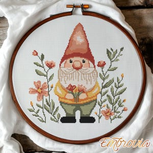 May include: Cross-stitch embroidery of a garden gnome holding flowers, surrounded by floral elements. The gnome wears a red hat, yellow jacket, and green trousers. The embroidery is set in a wooden hoop.