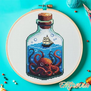 May include: Cross-stitch art of a ship in a bottle with an octopus. The design features a detailed illustration of a ship sailing on blue waves, with an orange octopus in the bottle. The bottle has a cork stopper and is set in a wooden embroidery hoop.