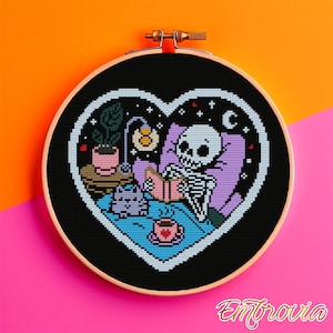 May include: A cross-stitch embroidery in a round wooden hoop. The design features a skeleton in bed reading, with a cat, a lamp, and a potted plant. The background is black, with a heart-shaped border. The color palette includes black, white, pink, purple, and blue.