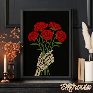 May include: A framed cross-stitch art piece featuring a bouquet of red flowers held by a skeletal hand against a black background. The flowers have green stems and leaves. The frame is black. The artwork is displayed on a dark surface.