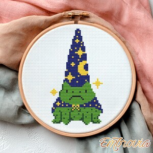 May include: A cross-stitch embroidery hoop featuring a green frog wearing a wizard hat and cape. The hat is dark blue with yellow stars and a crescent moon. The frog has a grumpy expression. The embroidery is set in a wooden hoop.