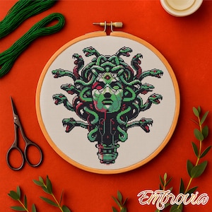 May include: A cross-stitch embroidery of Medusa in a wooden hoop. The design features a green face and snakes, with red accents. Green thread, scissors, and sprigs of greenery surround the hoop on an orange background. The word "Embrovia" is in the bottom right corner.
