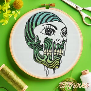 May include: Cross-stitch embroidery in a wooden hoop featuring a stylized skull design. The skull has green and black striped hair, with green and yellow dripping features. The eyes are detailed with black and white. The word "Embrovia" is in the bottom right corner.