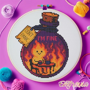 Peut inclure: Broderie au point de croix dans un cercle en bois. Le motif représente une bouteille de potion violette avec un personnage de flamme souriant à l'intérieur, entouré de flammes. Le texte comprend "DON'T PANIC" et "I'M FINE". Le fond est violet vif.