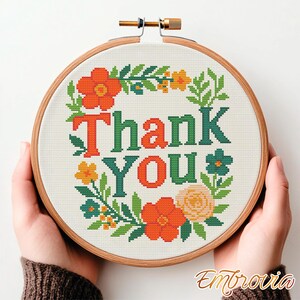 May include: A cross-stitch embroidery hoop with the words "Thank You" in green and orange, surrounded by a floral wreath. The design includes orange, yellow, and blue flowers with green leaves. The hoop is held by hands.