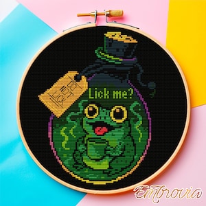 May include: Cross-stitch embroidery in a wooden hoop features a green frog with a cup, inside a black potion bottle. The bottle has a tag that says "Do Not Lick" and the text "Lick me?" is also present. The background is black.