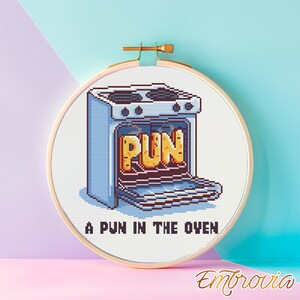 May include: Cross-stitch embroidery in a wooden hoop features a blue oven with the word "PUN" inside. The text "A PUN IN THE OVEN" is below the oven. The background is a gradient of blue and pink.