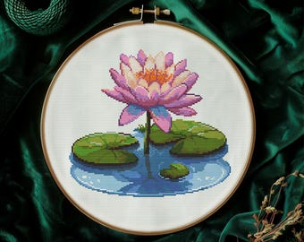 May include: A completed cross-stitch embroidery of a water lily in a wooden hoop. The flower has pink and orange petals, with green lily pads and blue water. The embroidery is set against a dark green background.