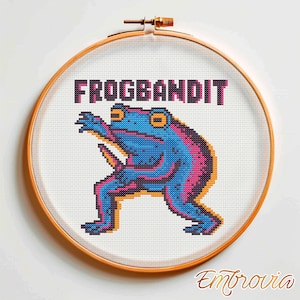May include: Cross-stitch embroidery in a wooden hoop featuring a blue frog with pink and orange accents. The text "FROGBANDIT" is above the frog. The embroidery is on white fabric. The artist's signature "Embrovia" is at the bottom.