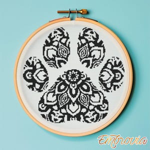 Può includere: Un telaio da ricamo a punto croce con un disegno di impronta di zampa in bianco e nero. L'impronta della zampa è dettagliata con motivi geometrici. Il telaio in legno è posto su uno sfondo azzurro. La parola "Embrovia" è scritta in corsivo.