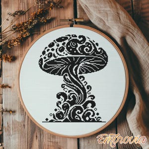 May include: A black and white cross-stitch mushroom design in a wooden embroidery hoop. The mushroom features intricate swirling patterns. The embroidery is set against a white fabric background. The word "Embrovia" is in the bottom right corner.