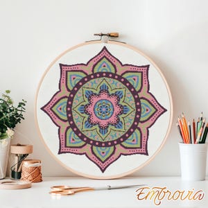 May include: A colorful mandala cross-stitch design in a wooden embroidery hoop. The design features pink, green, blue, and purple hues. The word "Embrovia" is written in gold script at the bottom right.