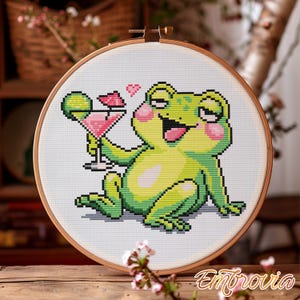May include: Cross-stitch embroidery of a cartoon frog holding a pink cocktail with a heart above it. The frog is green with pink cheeks and is set in a wooden embroidery hoop. The word "Embrovia" is written in cursive at the bottom.