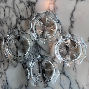 May include: Four clear glass coasters with a starburst design in the center. The coasters have a faceted, scalloped edge. The coasters are arranged on a marble surface.