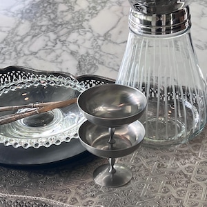 Op de afbeelding: Een zilverkleurig metalen dienblad met een ingewikkeld geometrisch patroon, met daarop een heldere glazen karaf met verticale witte strepen en een gepolijste zilveren rand, twee gestapelde zilverkleurige metalen schalen op een voet en een glazen bord.