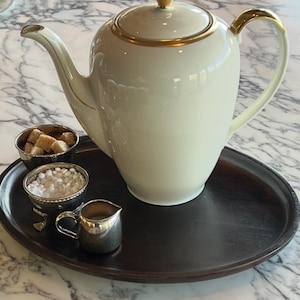 May include: A cream-colored teapot with gold trim and a matching lid sits on a dark wooden tray. Small silver bowls hold sugar cubes and a white granular substance, with a small silver creamer. The teapot has a curved handle and spout.