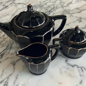 Può includere: Un set da tè di tre pezzi composto da una teiera, una lattiera e una zuccheriera con coperchi. Ogni pezzo è realizzato in ceramica nera lucida con lati sfaccettati distintivi e un sottile bordo color oro lungo i coperchi e i corpi.