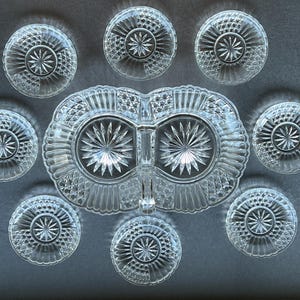 Op de afbeelding: Een serveerset van helder persglas bestaande uit één ovale verdeelde schaal met een centraal handvat en acht bijpassende kleine ronde bordjes. Elk stuk is voorzien van een gedetailleerd sterpatroon in het midden, omringd door een ruitvormig raster.