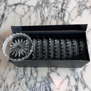 Op de afbeelding: Een set van heldere glazen onderzetters met een sterrenpatroon, gepresenteerd in een zwarte doos. De onderzetters hebben een diamantgeslepen patroon. De doos is open en toont de onderzetters op een marmeren oppervlak.